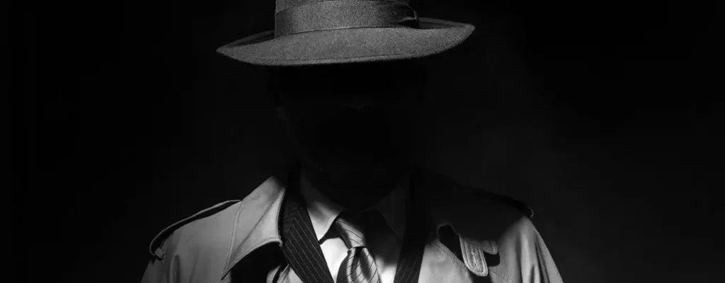 Shadowy black and white figure wearing a fedora and trench coat