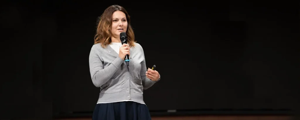 Woman with brown hair and wearing a gray sweater speaking into a microphone